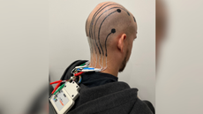 A man who is wearing a black sweatshirt is sat facing away from the camera. The e-tattoos can be seen across his scalp, with ink running down his head and joining up to wires at the base of his neck which are connected to a white device.